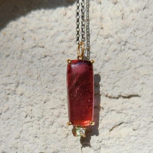 May include: A pendant necklace featuring a rectangular, deep red gemstone with gold accents. The pendant hangs from a silver chain and includes a small, light green gemstone at the bottom. The necklace is displayed against a textured, off-white background.