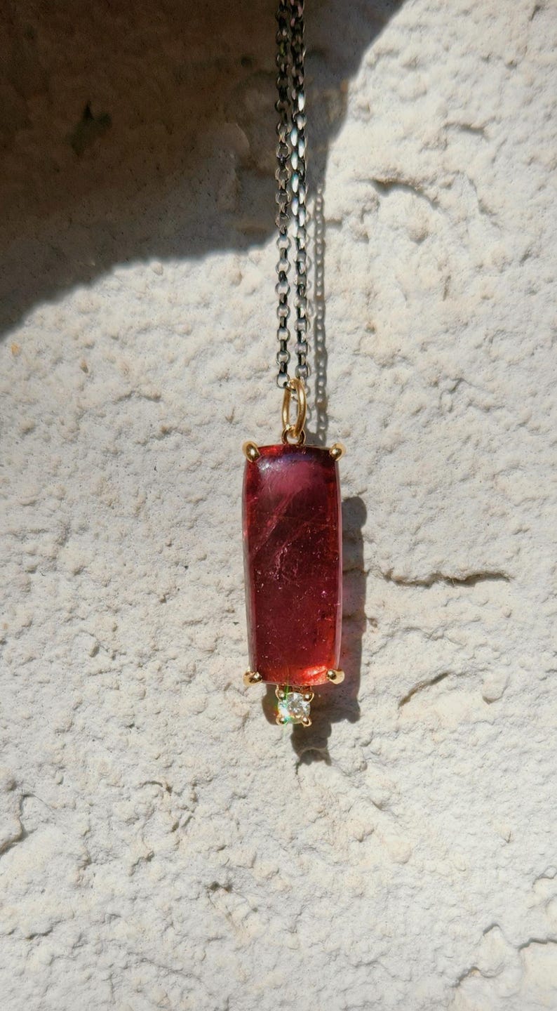May include: A pendant necklace featuring a rectangular, deep red gemstone with gold accents. The pendant hangs from a silver chain and includes a small, light green gemstone at the bottom. The necklace is displayed against a textured, off-white background.