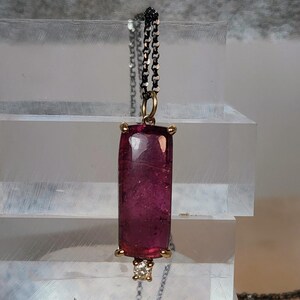 May include: A pendant necklace featuring a rectangular, deep pink gemstone. The gemstone is set in a gold-colored bezel with a small diamond accent at the bottom. The chain is a dark, linked design.