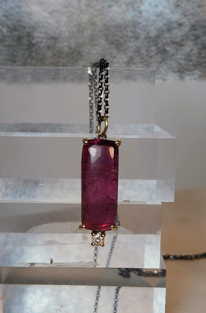 May include: A pendant necklace featuring a rectangular, deep pink gemstone. The gemstone is set in a gold-colored bezel with a small diamond accent at the bottom. The chain is a dark, linked design.