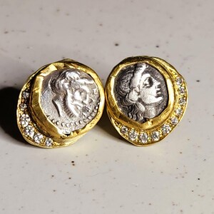 May include: A pair of gold earrings with a silver coin design. Each earring features a silver coin with a profile of a woman's face. The coins are surrounded by a gold frame with a row of small diamonds.