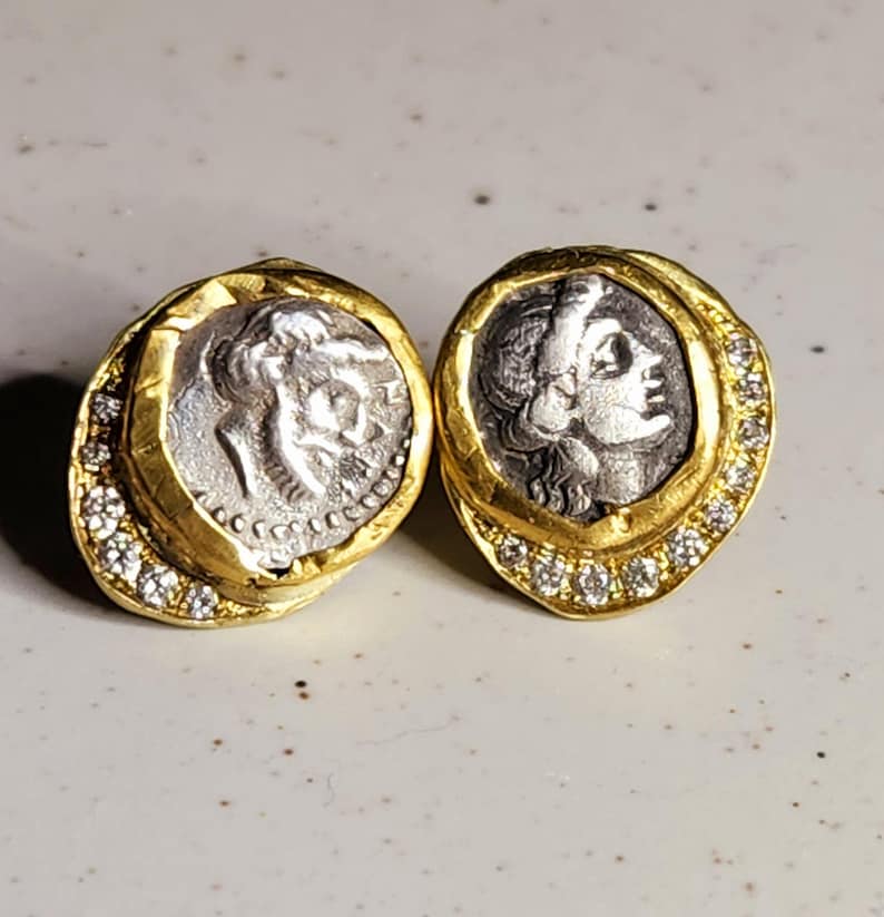 May include: A pair of gold earrings with a silver coin design. Each earring features a silver coin with a profile of a woman's face. The coins are surrounded by a gold frame with a row of small diamonds.
