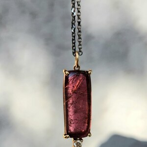 May include: A rectangular pendant necklace featuring a large, translucent, deep pink gemstone. The gemstone is set in a gold-colored frame and is suspended from a dark chain. A small, clear gemstone is set at the bottom of the pendant.