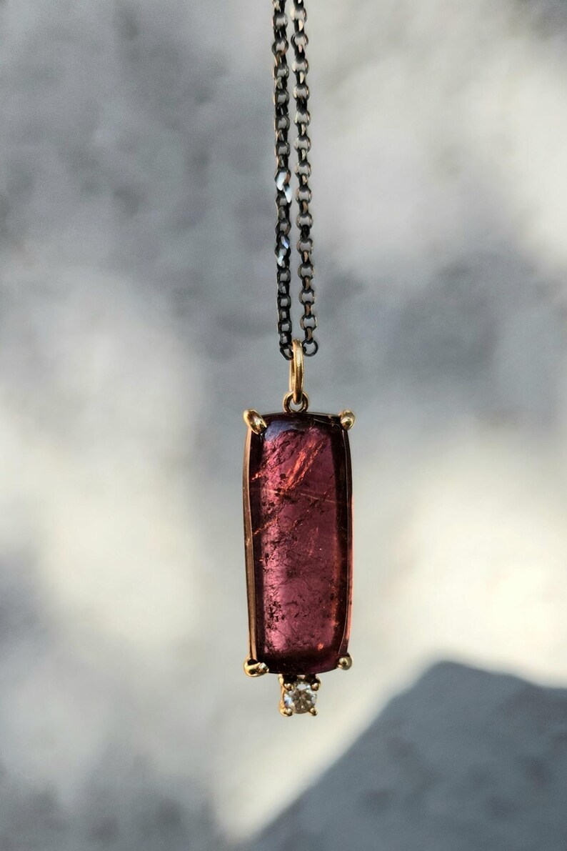 May include: A rectangular pendant necklace featuring a large, translucent, deep pink gemstone. The gemstone is set in a gold-colored frame and is suspended from a dark chain. A small, clear gemstone is set at the bottom of the pendant.
