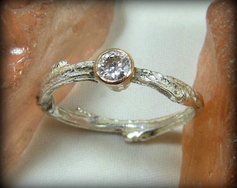 May include: A silver ring with a twig-like band and a gold bezel setting holding a round, clear gemstone. The ring's design evokes a natural, organic aesthetic, perfect for everyday wear.
