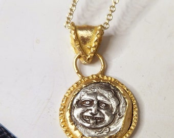 May include: A gold chain necklace with a pendant featuring a silver face with a toothy grin. The pendant is surrounded by a gold rim with a textured design.