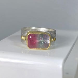 May include: A silver ring featuring a rectangular watermelon tourmaline gemstone set in a gold bezel. The gemstone displays pink and green hues. The ring has a textured band and small gold accents.
