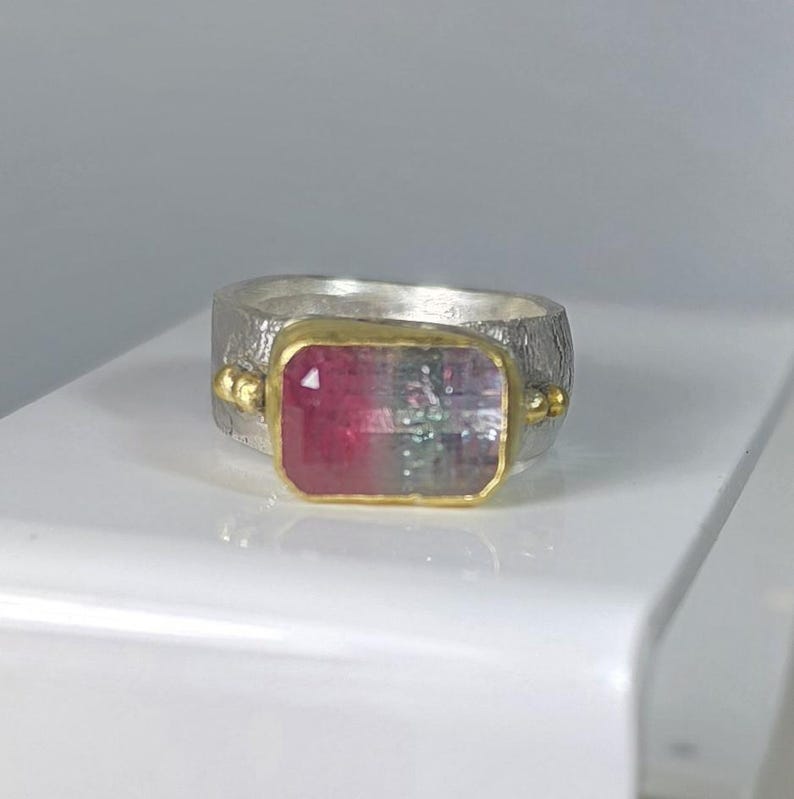May include: A silver ring featuring a rectangular watermelon tourmaline gemstone set in a gold bezel. The gemstone displays pink and green hues. The ring has a textured band and small gold accents.