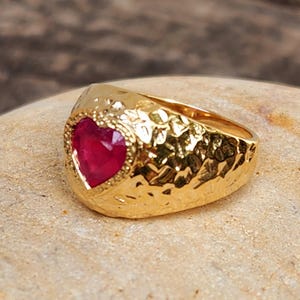 Peut inclure: Une bague dorée avec une texture martelée. La bague présente une pierre précieuse rouge foncé en forme de cœur. La pierre est entourée d'une bordure texturée, accentuant le design. La bague est posée sur une surface brun clair.