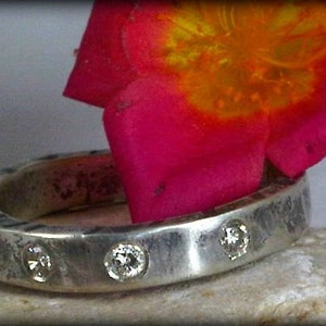 Pode incluir: Um anel de prata com três pequenos diamantes cravejados na banda. O anel é texturizado e tem um visual rústico. O anel está apoiado em uma superfície branca com uma flor rosa atrás.