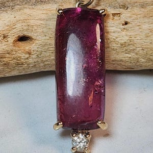 May include: A rectangular, deep magenta pendant with a gold setting and a small, square diamond at the bottom. The pendant is suspended from a delicate silver chain. The gemstone has a polished, reflective surface.