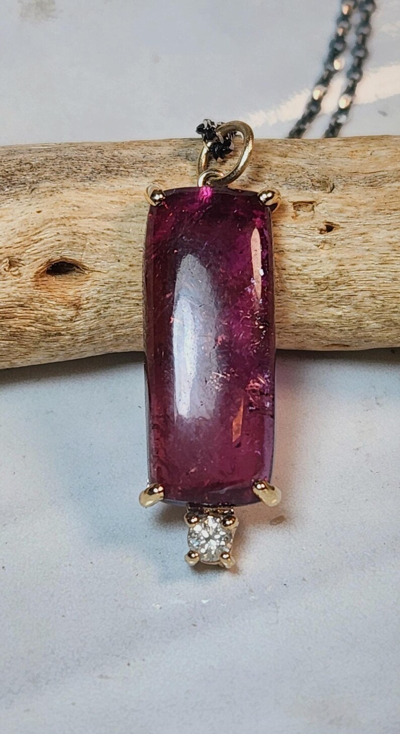 May include: A rectangular, deep magenta pendant with a gold setting and a small, square diamond at the bottom. The pendant is suspended from a delicate silver chain. The gemstone has a polished, reflective surface.