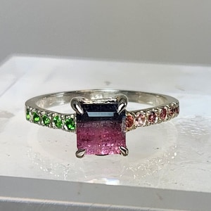May include: A silver ring with a large, rectangular pink gemstone and a dark blue top. The band is decorated with a gradient of small gemstones, changing from green to pink. The ring is set against a white background.