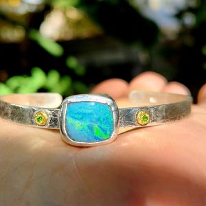 May include: Silver cuff bracelet featuring a large, square, iridescent blue opal center stone. The bracelet has a textured finish and is accented with two small, round, gold-colored settings with green gemstones.