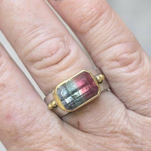 May include: A silver ring featuring a rectangular watermelon tourmaline gemstone set in a gold bezel. The gemstone shows a gradient of pink, green, and clear facets. Small gold beads adorn the ring's band.
