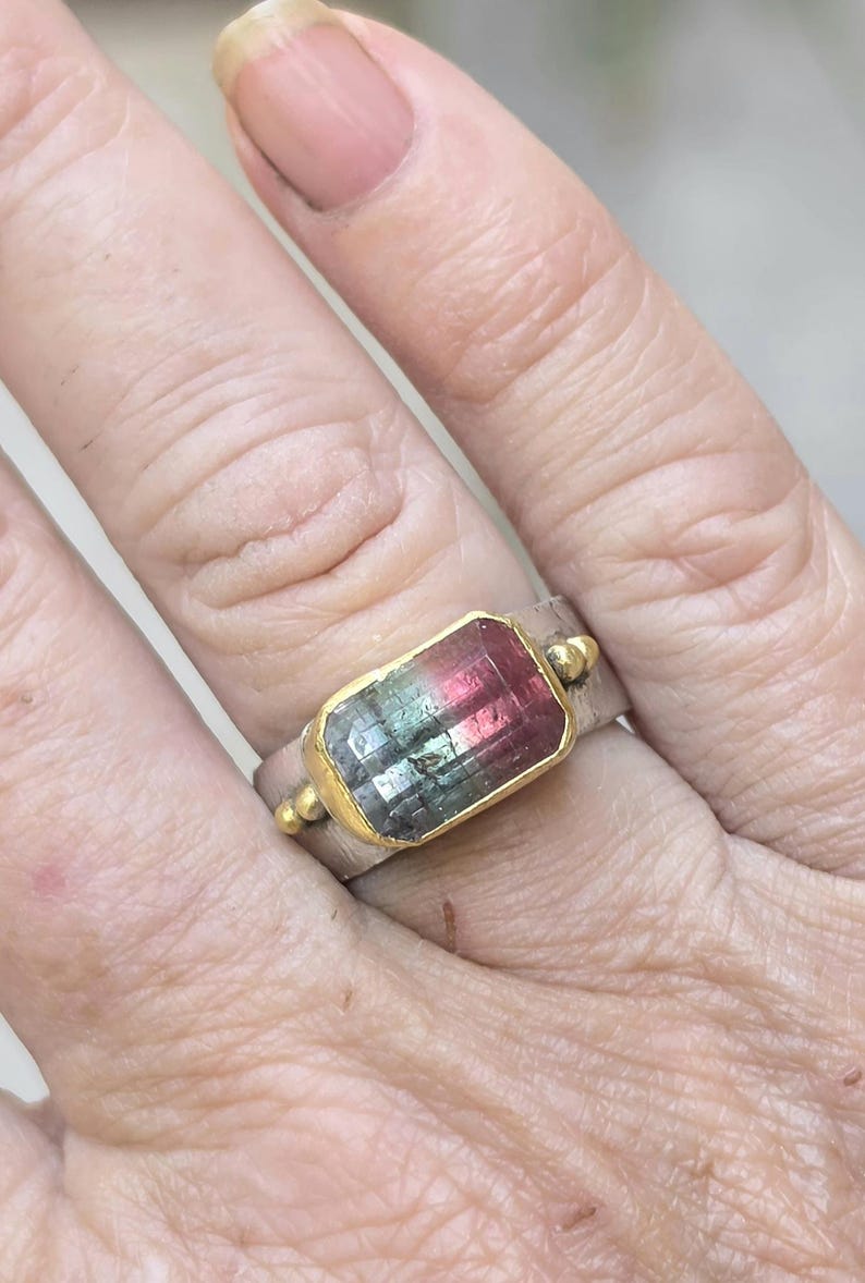 May include: A silver ring featuring a rectangular watermelon tourmaline gemstone set in a gold bezel. The gemstone shows a gradient of pink, green, and clear facets. Small gold beads adorn the ring's band.