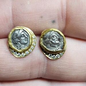 May include: A pair of gold earrings with a silver coin design. Each earring features a silver coin with a profile of a woman's face, surrounded by a gold rim and a row of small diamonds.