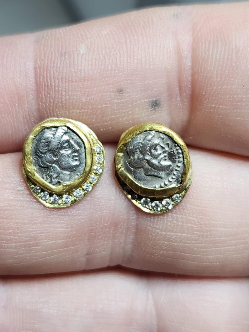 May include: A pair of gold earrings with a silver coin design. Each earring features a silver coin with a profile of a woman's face, surrounded by a gold rim and a row of small diamonds.