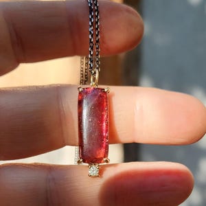 May include: A rectangular, red gemstone pendant necklace. The gemstone is set in a gold-colored frame and hangs from a delicate silver chain. A small, clear stone is set below the gemstone. The necklace is held between fingers.