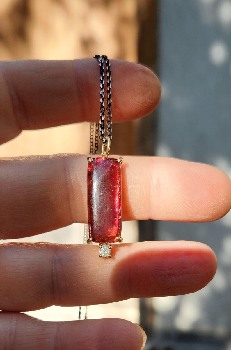 May include: A rectangular, red gemstone pendant necklace. The gemstone is set in a gold-colored frame and hangs from a delicate silver chain. A small, clear stone is set below the gemstone. The necklace is held between fingers.