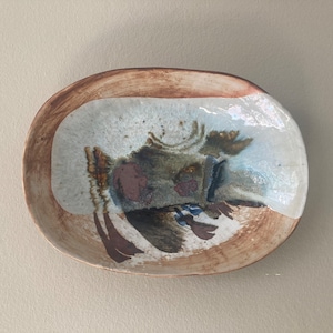 May include: An oval ceramic dish with a brown rim and a white interior. The dish has a unique abstract design in shades of brown, green, and white.