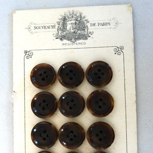 May include: A card with 12 brown buttons arranged in a grid. The card has the text "Nouveauté de Paris" and "Registered" printed on it. The card also has the text "Superior Quality" printed on it.