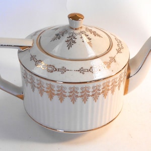 May include: White teapot with gold trim and a decorative floral pattern. The teapot has a lid and a handle.