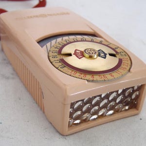 May include: A pink vintage light meter with a rotating dial that has markings for film speed and shutter speed. The dial has a gold center with the General Electric logo. The light meter has a clear plastic cover over the light sensor.