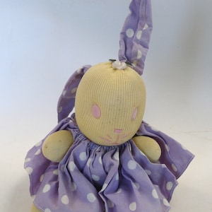 May include: A handmade sock bunny doll wearing a purple dress with white polka dots. The bunny has a long, pointed ear and pink embroidered eyes.