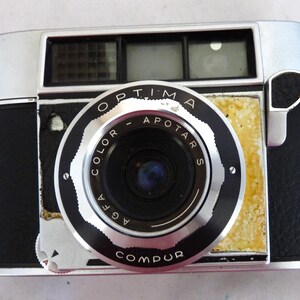 May include: A black and silver 35mm film camera with a silver lens labeled "Agfa Color-Apotars" and "Compur". The camera has a black leather covering and a silver top with a viewfinder.