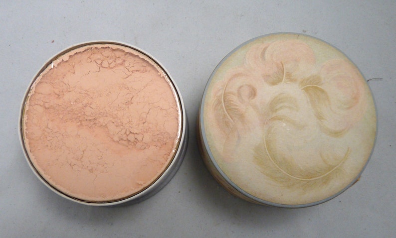 Peut inclure: Un compact rond blanc avec un motif de plumes sur le dessus. Le fond du compact est ouvert, r&eacute;v&eacute;lant une poudre de couleur p&ecirc;che clair.