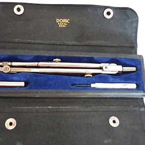 May include: A vintage drafting compass set in a black leather case with a blue velvet interior. The set includes a silver-colored compass, a small silver tool, and a silver pencil holder. The case is marked "DORIC K&E Co. 8004".