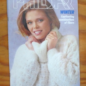 May include: Magazine cover featuring a woman wearing a cream-colored, textured knit sweater with a turtleneck. The magazine title is "PHILDAR MAILLES" with the word "WINTER" and the tagline "Captivating combinations of fibers". The magazine is on a wooden surface.