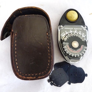May include: A vintage Norwood Director light meter with a leather case and several black aperture discs. The meter has a black and clear face with white and red markings. The leather case is brown with visible stitching.