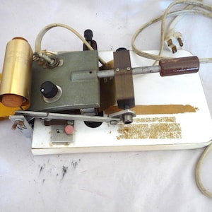May include: A vintage gold and silver foil stamping machine with a white base and a brown handle. The machine has a gold roller and a silver lever. The machine is plugged in and ready to use.