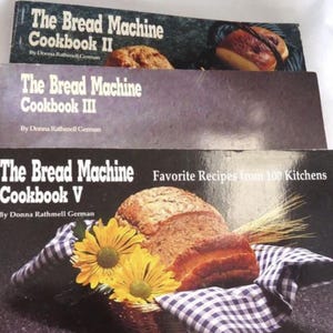 May include: Three vintage cookbooks titled "The Bread Machine Cookbook" are stacked. The covers feature images of bread loaves. The titles are "II", "III", and "V". The author's name, Donna Rathmell German, is visible on each book.