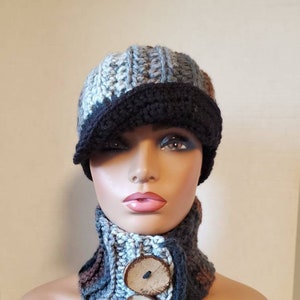 May include: A crocheted hat and neck warmer set in shades of blue, gray, and brown. The hat has a black brim and the neck warmer has two large wooden buttons.