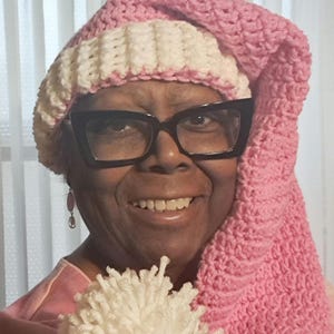 May include: A pink and white crocheted hat with a long, pointed end. The hat is worn by a person wearing black-framed glasses. A white yarn pom-pom is held in front of the person.