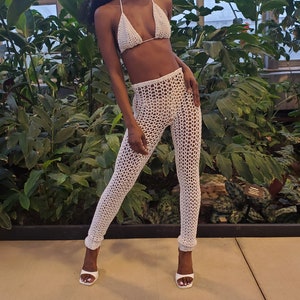 May include: A white crochet bikini top and matching pants. The model is wearing white heels and is standing in a greenhouse.