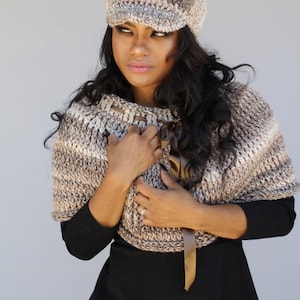 May include: A woman wearing a brown and beige crocheted hat with a brim and a matching brown and beige crocheted shawl. She is wearing a black long-sleeved shirt and blue jeans.