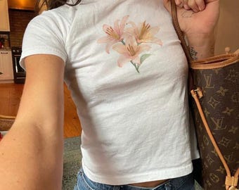 May include: A white t-shirt with a floral print of pink lilies. The shirt is cropped and has short sleeves.