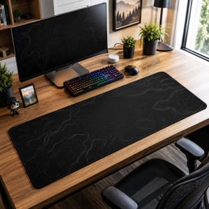 Peut inclure: Un grand tapis de bureau rectangulaire présentant une surface noire avec de fines lignes de contour topographiques claires. Le tapis est placé sur un bureau en bois devant un écran d'ordinateur, un clavier mécanique et une souris.