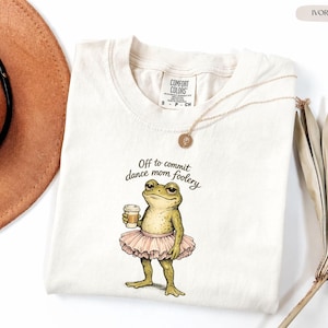 Puede incluir: Camiseta color marfil con una rana con tutú sosteniendo una taza de café. El texto "Off to commit dance mom foolery" está impreso en la camiseta. Un collar dorado está drapeado sobre la camiseta.
