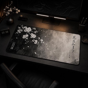 Puede incluir: Una alfombrilla de escritorio rectangular con un fondo degradado en negro y gris y una ilustración de una rama de cerezo en flor blanca. El diseño incluye pétalos dispersos y caligrafía china negra vertical en el lado derecho.