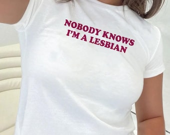 Op de afbeelding: Witte crop top met korte mouwen. De tekst "NOBODY KNOWS I'M A LESBIAN" is in donkerroze op de voorkant van de shirt gedrukt. Het shirt is gemaakt van een zacht, lichtgewicht materiaal.