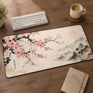 May include: A rectangular desk mat featuring a printed illustration of pink cherry blossoms on a dark branch in the foreground, with a pale landscape of mountains, trees, and a sun in the background. The mat includes black Japanese text.