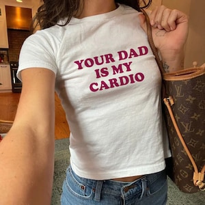 Puede incluir: Camiseta blanca recortada con el texto "YOUR DAD IS MY CARDIO" impreso en rosa.