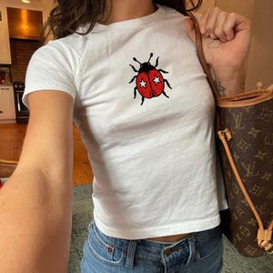 以下が含まれることがあります： 白い半袖Tシャツで、中央に黒い斑点と各羽に白い星が描かれた赤いテントウムシのグラフィックがプリントされています。青いデニムジーンズと、模様入りの茶色のショルダーバッグを合わせています。
