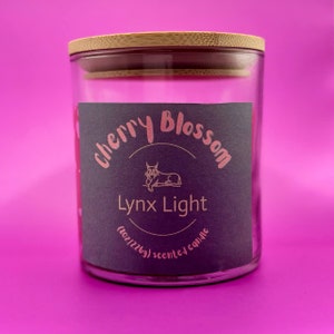 May include: A glass jar candle with a black label that says "Cherry Blossom Lynx Light (8oz/226g) scented candle". The label has a pink stylized illustration of a lynx.