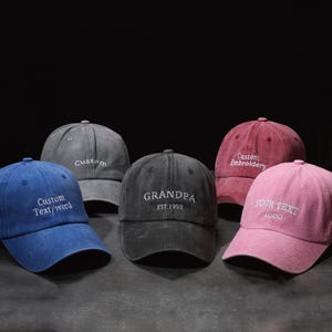 Peut inclure: Cinq casquettes de baseball de couleurs variées : bleu, gris, noir, bordeaux et rose. Chaque casquette présente un texte ou une broderie personnalisée en blanc. La casquette noire indique "GRANDPA EST.1998". La rose dit "YOUR TEXT LOGO".
